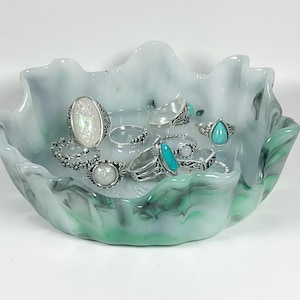 May include: A shallow, wavy-edged bowl made of white glass with swirling green and grey marbled patterns. Inside the bowl are several silver-toned rings, including pieces with iridescent white stones, turquoise-coloured teardrops, and floral designs.
