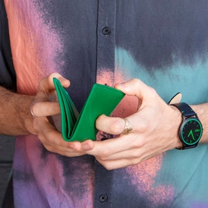 Pode incluir: Uma pessoa est&aacute; segurando uma carteira de couro verde em suas m&atilde;os. A carteira est&aacute; dobrada ao meio e a pessoa est&aacute; segurando-a aberta. A pessoa est&aacute; vestindo uma camisa colorida e um rel&oacute;gio preto com um mostrador verde.