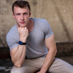 Puede incluir: Un hombre con una camiseta gris y pantalones caqui se sienta con el brazo apoyado en la rodilla. Lleva un reloj de pulsera azul y negro.