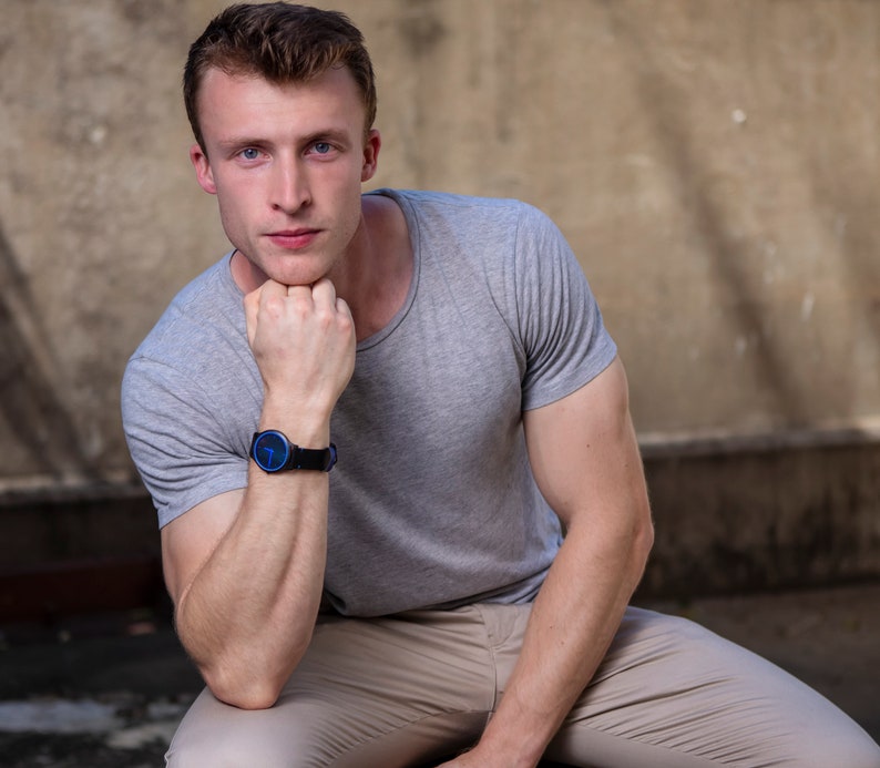 Puede incluir: Un hombre con una camiseta gris y pantalones caqui se sienta con el brazo apoyado en la rodilla. Lleva un reloj de pulsera azul y negro.