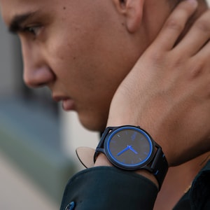 Puede incluir: Un reloj de pulsera negro con una esfera azul y agujas azules. El reloj tiene una correa de cuero negro.