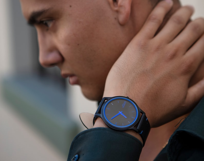 Puede incluir: Un reloj de pulsera negro con una esfera azul y agujas azules. El reloj tiene una correa de cuero negro.