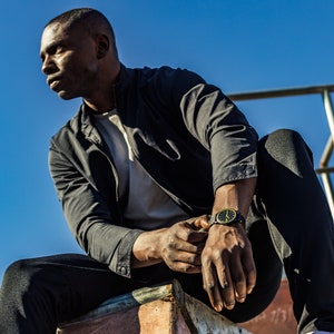 May include: A man wearing a black jacket, white t-shirt, and black trousers sits on a concrete ledge. He is wearing a black watch with a yellow face.
