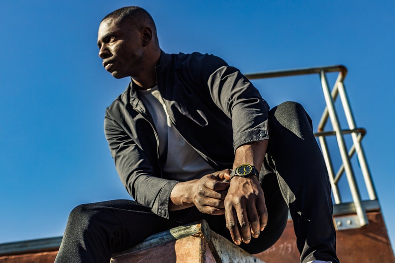 May include: A man wearing a black jacket, white t-shirt, and black trousers sits on a concrete ledge. He is wearing a black watch with a yellow face.
