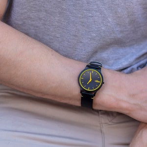 May include: A black wristwatch with a yellow rim around the face and a black leather strap. The watch face has black hands and a small white logo that says "Kido".