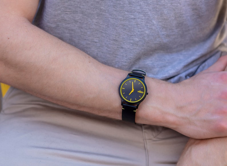 May include: A black wristwatch with a yellow rim around the face and a black leather strap. The watch face has black hands and a small white logo that says "Kido".