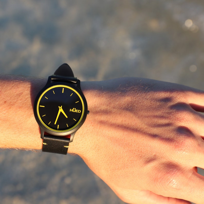 May include: A black wristwatch with a yellow face and black leather strap. The watch face has yellow hour and minute hands and the word "HUED" printed in yellow.