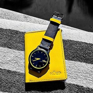 May include: A black wristwatch with a yellow face and black leather strap is sitting on a yellow leather wallet with the word "HUED" printed on it.