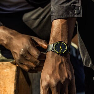 May include: A black wristwatch with a yellow face and black leather strap. The watch has a simple design with a black background and yellow hour and minute hands.