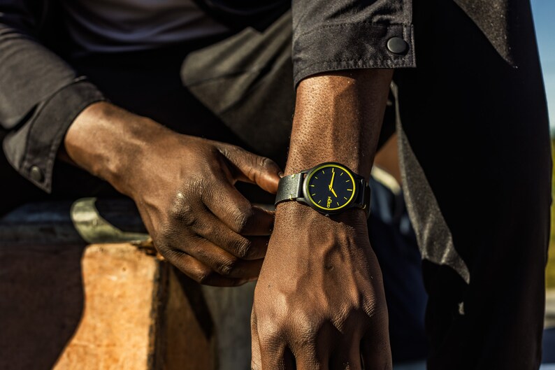 May include: A black wristwatch with a yellow face and black leather strap. The watch has a simple design with a black background and yellow hour and minute hands.