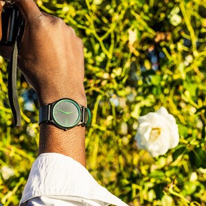 Pode incluir: Uma pessoa usando um rel&oacute;gio preto com um mostrador verde e uma pulseira preta. O rel&oacute;gio est&aacute; no pulso esquerdo. A pessoa est&aacute; usando uma camisa branca e uma jaqueta azul. O fundo &eacute; uma imagem desfocada de folhas verdes e flores brancas.