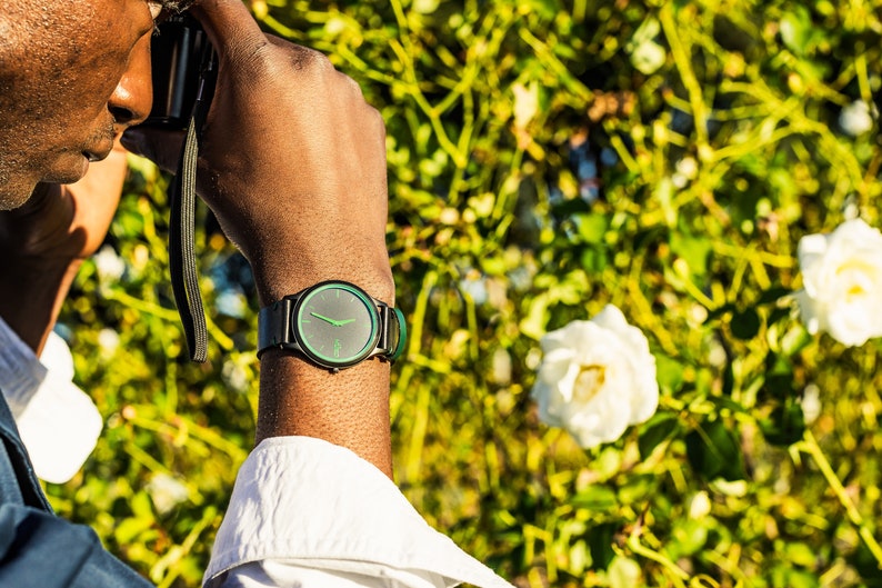 Pode incluir: Uma pessoa usando um rel&oacute;gio preto com um mostrador verde e uma pulseira preta. O rel&oacute;gio est&aacute; no pulso esquerdo. A pessoa est&aacute; usando uma camisa branca e uma jaqueta azul. O fundo &eacute; uma imagem desfocada de folhas verdes e flores brancas.