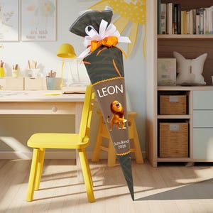 May include: A brown and green school cone with the name "LEON" in white. A cartoon lion is printed below the name. The cone is decorated with a white and orange bow. The text "Schulkind 2026" is printed on the cone.