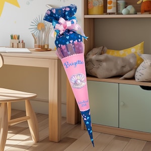 Può includere: Un cono scolastico rosa e blu con un design a conchiglia e il nome "Brigitte". Il cono è decorato con cuori e un fiocco. La parte inferiore del cono riporta la scritta "Schulkind 2026".