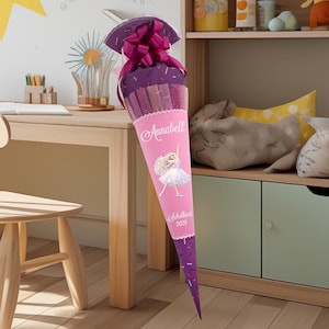 Könnte beinhalten: Eine rosa-lila Schultüte mit dem Namen "Annabell" und einer Ballerina-Illustration. Die Tüte ist mit einer großen lila Schleife verziert und mit Süßigkeiten gefüllt. Der Text "Schulkind 2026" ist ebenfalls sichtbar.