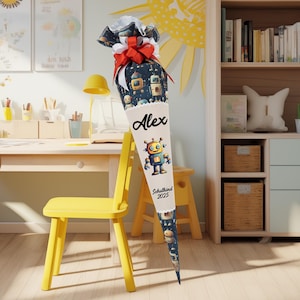 Op de afbeelding: Een crèmekleurige, kegelvormige schooltas met een robotontwerp en de naam "Alex". De tas is versierd met een marineblauwe stof aan de bovenkant, een rode strik en de tekst "Schulkind 2025."