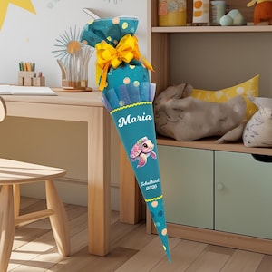 Peut inclure: Un cornet scolaire bleu sarcelle et jaune avec une illustration de tortue de dessin animé. Le cône est décoré de pois et d'un ruban jaune. Le nom "Maria" et l'année "2026" sont imprimés.