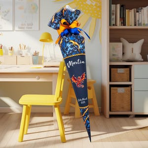 Op de afbeelding: Een marineblauwe schooltuit met een drakenontwerp en de naam "Martin" in witte letters. De tuit is versierd met blauwe en oranje linten en een drakenillustratie. De tekst "Schulkind 2020" is ook zichtbaar.