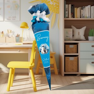 Könnte beinhalten: Ein blauer Schultüte mit einem weißen Eisbären-Motiv. Die Tüte ist mit weißer Spitze und einer weißen Schleife verziert. Der Text "Barney" und "Schulland 2025" ist auf die Tüte gedruckt.