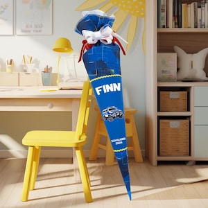 Op de afbeelding: Een blauwe en witte schooltuit met de naam "Finn" in witte blokletters. De tuit heeft een politieauto-afbeelding en de tekst "SCHULKIND 2026". Hij is vastgebonden met een witte strik en rode linten.