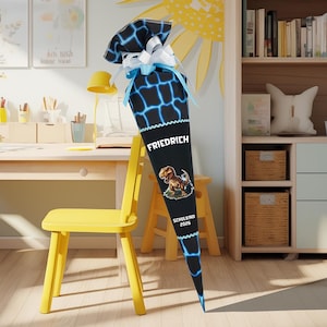 Könnte beinhalten: Eine schwarze und blaue Schultüte mit einem Dinosaurier-Motiv und dem Namen "FRIEDRICH" in Weiß. Sie ist mit weißen und blauen Bändern verziert. Der Text "SCHULKIND 2026" ist ebenfalls sichtbar.
