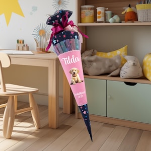 May include: A pink and blue school cone with a picture of a golden retriever puppy. The cone has the name "Tilda" and the text "Schulkind 2023" on it.
