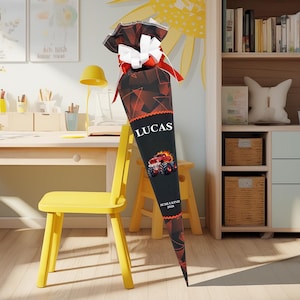 May include: A large, cone-shaped school bag with a black base and a red and black geometric pattern. The bag features the name "LUCAS" in white, a monster truck graphic, and the text "SCHULKIND 2026". It is topped with a red and white bow.