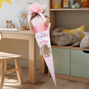 Könnte beinhalten: Ein rosa-weißer Schultüte mit einem Pferde-Motiv und dem Namen "Emilia" darauf. Die Tüte ist mit Blumen und einem Band verziert.