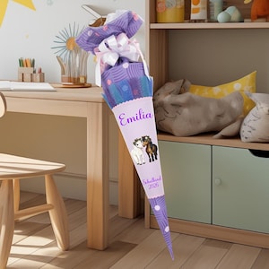 May include: A large, cone-shaped school bag in shades of purple, blue, and white, decorated with a bow and the name "Emilia." The bag features illustrations of two horses and the text "Schulkind 2026."