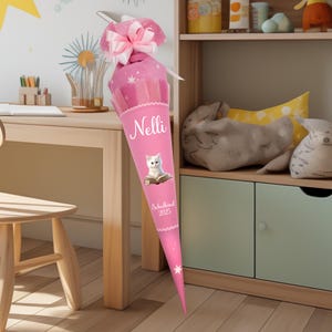 Può includere: Cono scolastico rosa con un gatto bianco che legge un libro, stelle e il nome "Nelli" e il testo "Schulanfang 2025".