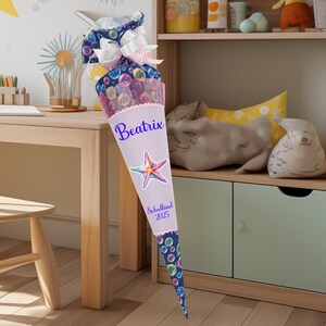 Könnte beinhalten: Ein rosa-blauer Schultüte mit einem weißen Etikett. Das Etikett trägt den Namen "Beatrix" und den Text "Schulkind 2025". Die Tüte ist mit einem bunten Seestern und einem Muster aus Blasen verziert.