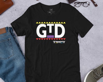 May include: Black t-shirt with a white graphic that says "GTD" with a purple line and the word "TOMMY" underneath. The graphic is surrounded by a colorful pattern of triangles.