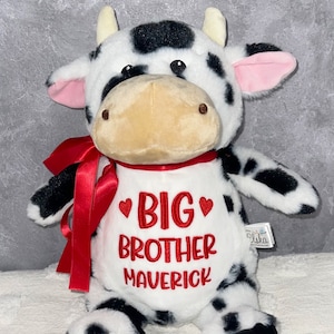 May include: A plush cow toy with black spots, white fur, and a red bow. The cow has a white bib embroidered with "BIG BROTHER MAVERICK" in red. The toy has a tan snout and pink inner ears.