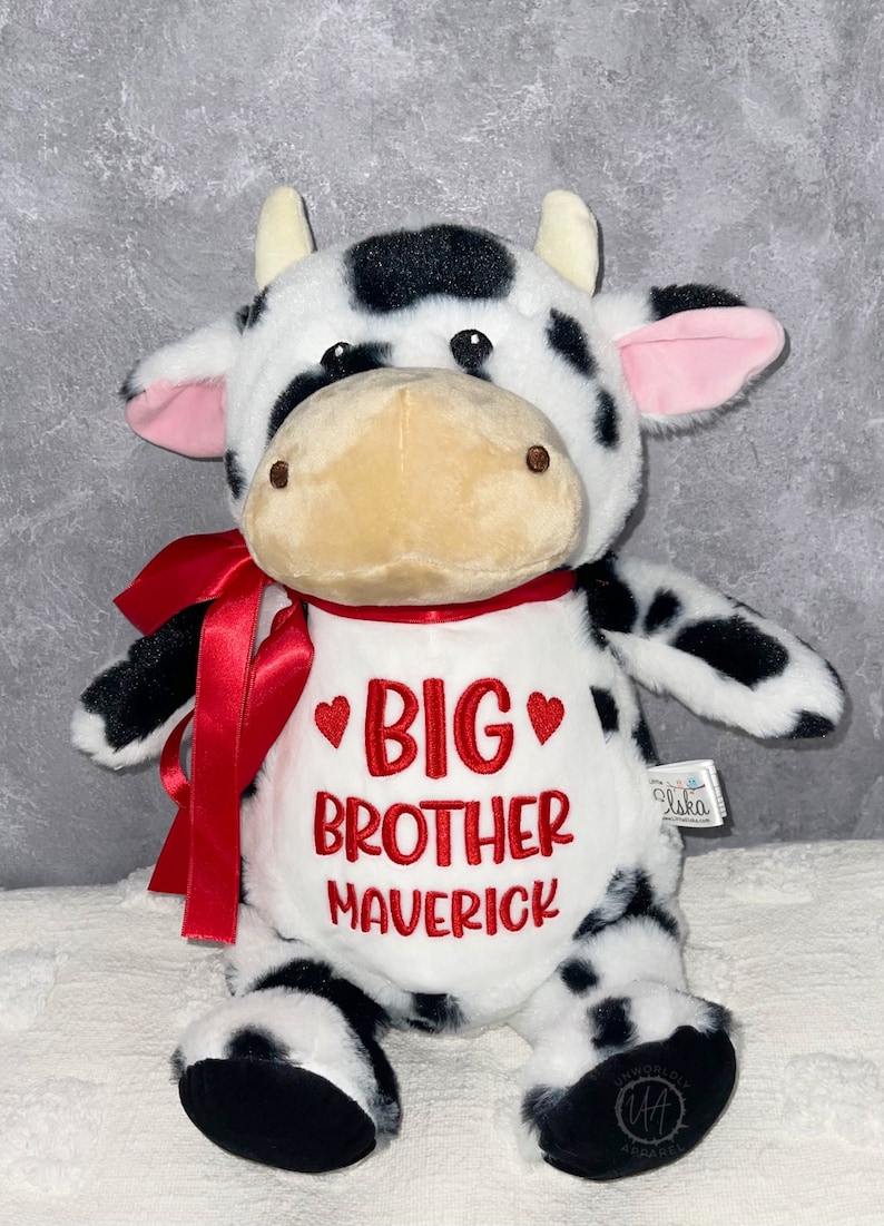 May include: A plush cow toy with black spots, white fur, and a red bow. The cow has a white bib embroidered with "BIG BROTHER MAVERICK" in red. The toy has a tan snout and pink inner ears.