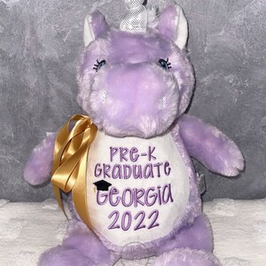 May include: A plush purple unicorn with a white and black polka-dot horn. The unicorn has embroidered text that reads "Pre-K Graduate Georgia 2022". A gold ribbon is tied around its neck.