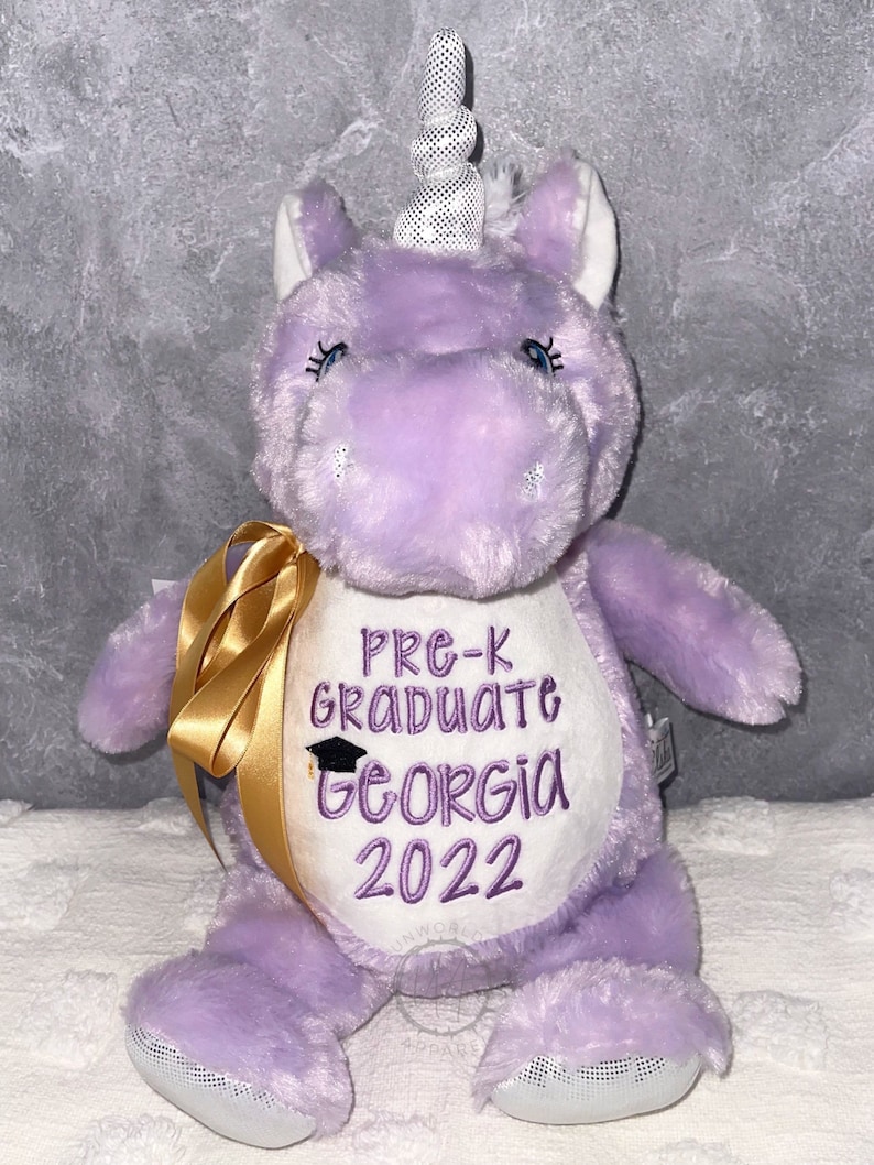 May include: A plush purple unicorn with a white and black polka-dot horn. The unicorn has embroidered text that reads "Pre-K Graduate Georgia 2022". A gold ribbon is tied around its neck.