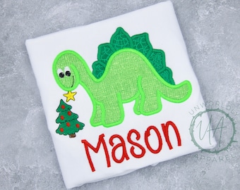 May include: White t-shirt featuring a green dinosaur holding a star and a Christmas tree. The name "Mason" is embroidered in red. The design is outlined in green.