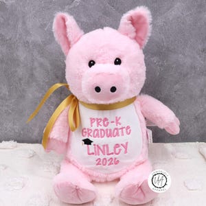 May include: A pink plush pig wearing a white bib embroidered with "Pre-K Graduate Linley 2026" in pink script. The pig has black eyes, a black snout, and a gold ribbon. The pig is a graduation gift.