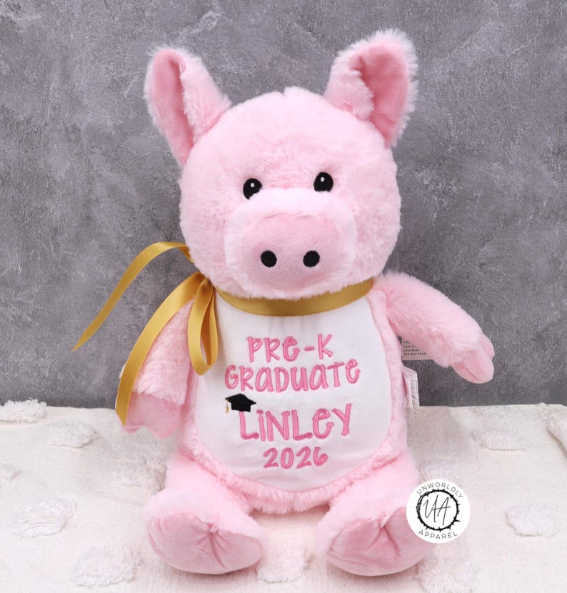 May include: A pink plush pig wearing a white bib embroidered with "Pre-K Graduate Linley 2026" in pink script. The pig has black eyes, a black snout, and a gold ribbon. The pig is a graduation gift.