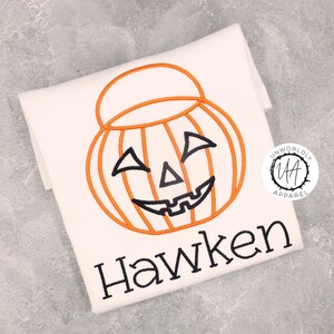 Puede incluir: Camiseta blanca con un diseño bordado de calabaza naranja, cara y asa negras. El nombre "Hawken" está bordado en negro debajo de la calabaza. La camiseta está doblada. Prenda con temática de Halloween.