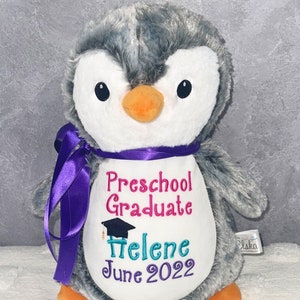 May include: A plush penguin toy with gray and white fur, an orange beak, and black eyes. It has a purple ribbon around its neck and embroidered text that reads "Preschool Graduate", a name, and "June 2022".