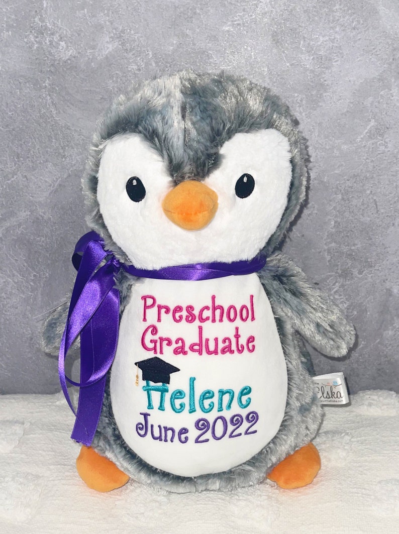 May include: A plush penguin toy with gray and white fur, an orange beak, and black eyes. It has a purple ribbon around its neck and embroidered text that reads "Preschool Graduate", a name, and "June 2022".