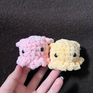 Puede incluir: Dos pequeños juguetes de pulpo de ganchillo. Uno es rosa claro, el otro es amarillo. Ambos tienen ojos de botón negros y una forma redondeada. Los juguetes se sostienen en una mano sobre un fondo marrón oscuro.