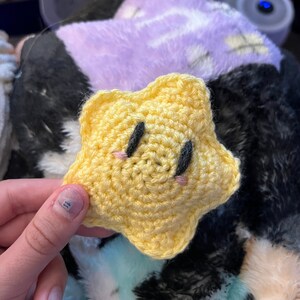 May include: A handmade, yellow crochet star with black eyes and pink cheeks. The star is a plush toy, likely for a child. The star is held in a person's hand, with a soft, multi-colored blanket in the background.