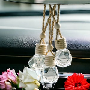 May include: Five clear glass car air fresheners hanging from a rearview mirror in a car. The air fresheners are shaped like diamonds and have wooden tops. The rearview mirror has the text "Filtered Finesse" written on it.