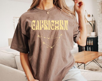 Könnte beinhalten: Ein braunes T-Shirt mit einer gelben Grafik des Sternzeichen Steinbock und dem Wort "Steinbock" in gelben Buchstaben.