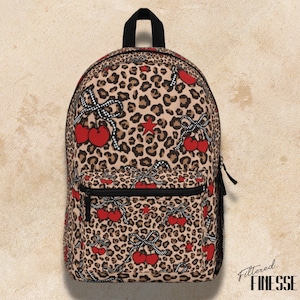 Pode incluir: Uma mochila com estampa de leopardo, adornada com cerejas vermelhas, laços xadrez brancos e pretos e uma estrela vermelha. A mochila tem um zíper preto, um bolso frontal e uma alça preta. O texto "Filtered Finesse" está no canto inferior direito.