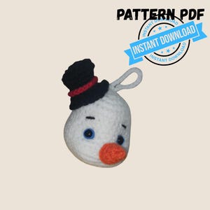 May include: A white crocheted snowman head ornament with a black top hat featuring a red band, blue eyes, and an orange nose. The item includes a hanging loop and text that reads PATTERN PDF INSTANT DOWNLOAD in the upper right corner.