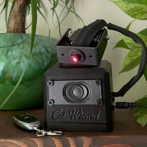 May include: A black electronic device labelled Cat Wizard with a central speaker, a top-mounted rotating component featuring a glowing red light, and a black strap. A small silver remote control with a metal clip is positioned in front of the unit.