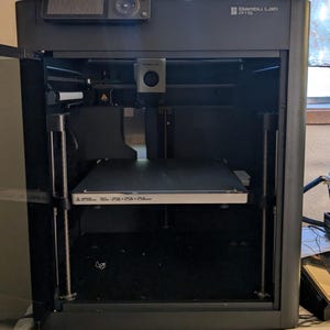 May include: A black 3D printer with a clear door open, revealing the print bed and a silver metal plate. The printer has a black and silver control panel on the top and the text "Bambu Lab P1S" on the front.